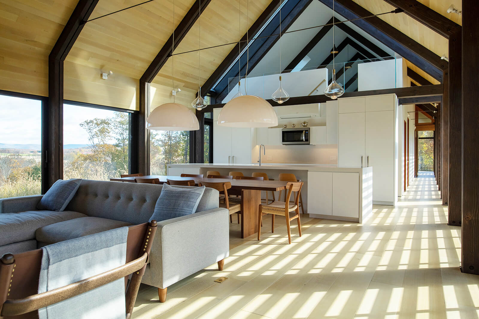 Modern open-concept living and dining area with high ceilings, exposed beams, large windows, a gray sofa, wooden dining table, and a bright, airy kitchen with white cabinetry and pendant lights. Sunlight streams across the floor.