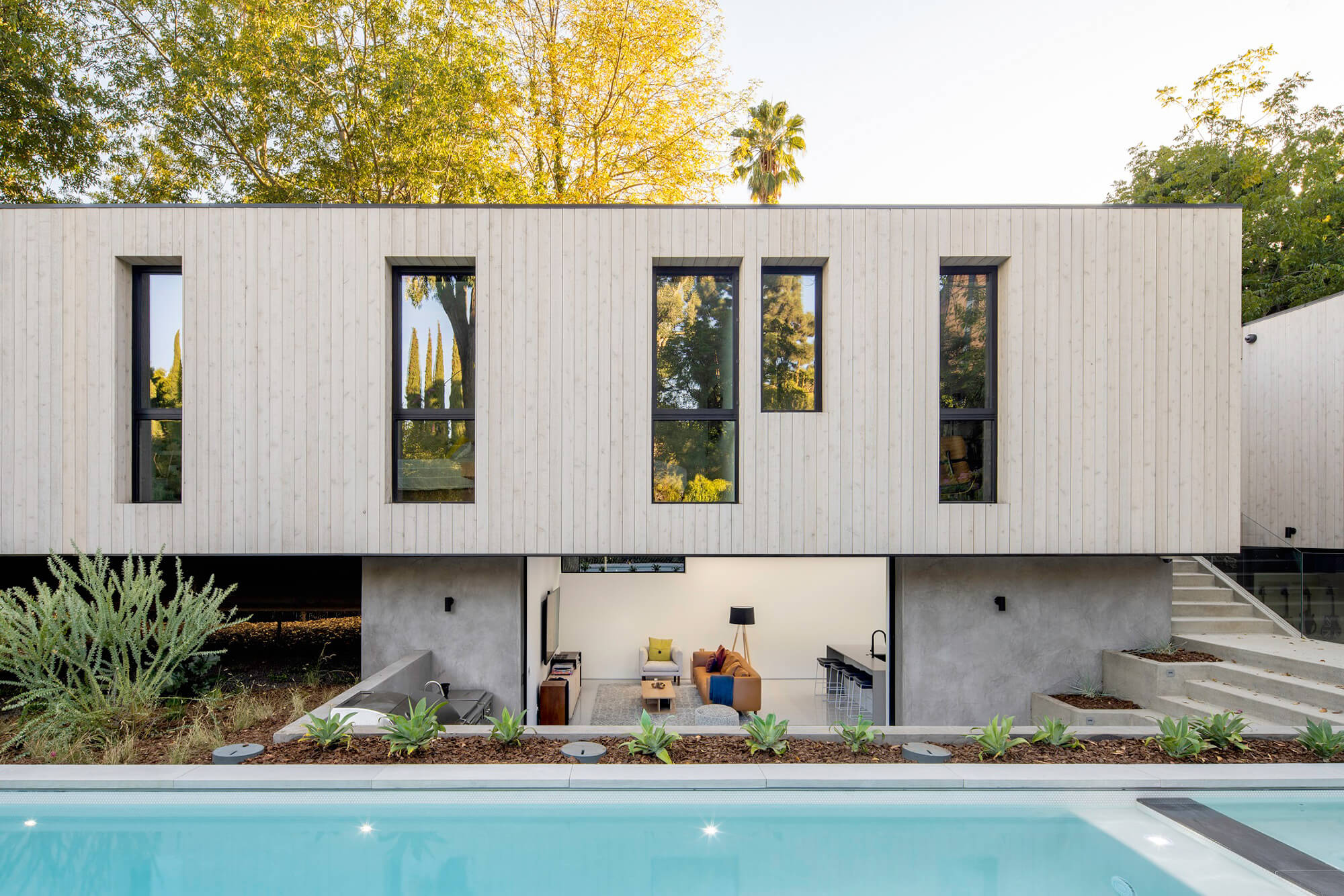 Modern two-story house with a light wood exterior, large vertical windows, and an open lower level facing a swimming pool. The landscaped yard features greenery and trees in the background.