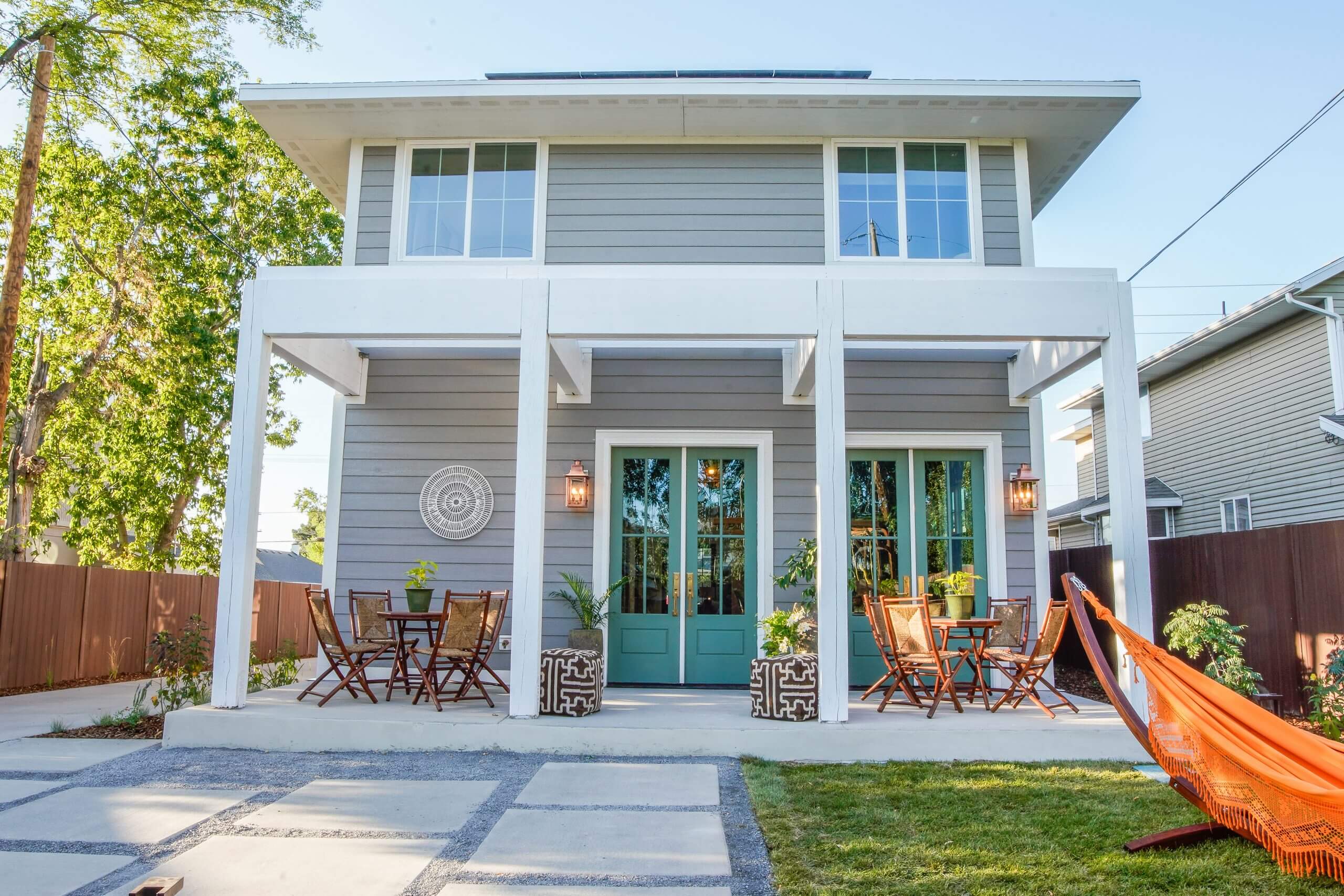 A modern two-story gray house with teal double doors, a covered patio, outdoor dining sets, potted plants, and a bright orange hammock on a green lawn. The yard features concrete pavers and is fenced with wood.