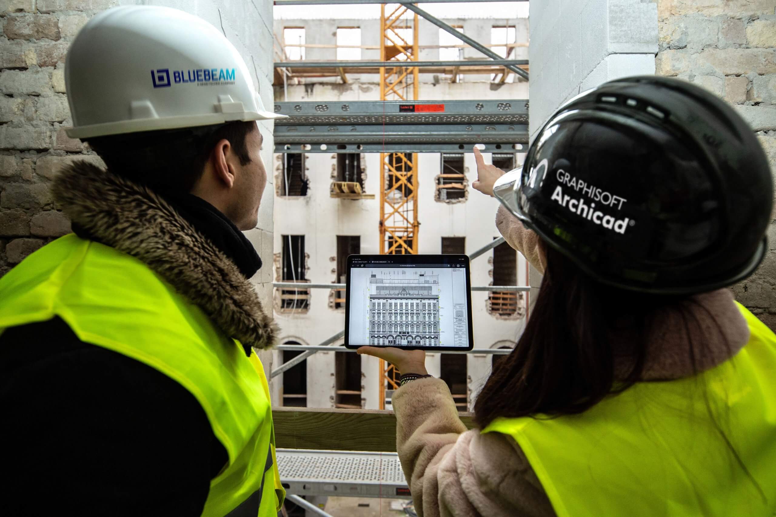 Two construction workers in hard hats and high-visibility vests review digital building plans on a tablet, utilizing BIM technology, at a construction site. A building facade and crane are visible in the background as one worker points towards the structure.