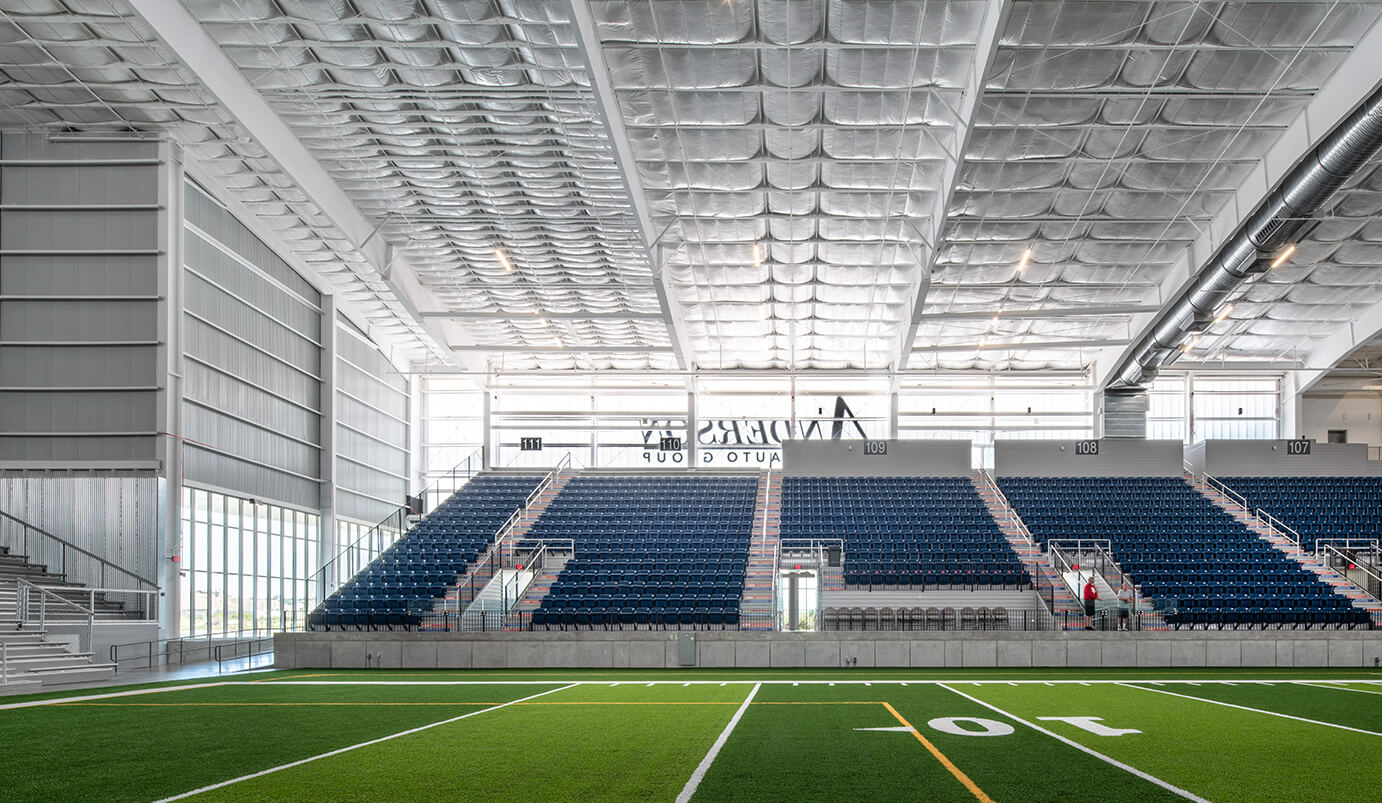 Interior view of a modern indoor sports stadium featuring blue bleachers, large windows, bright lighting, and artificial turf marked with white yard lines. An “Ander’s Auto Group” sign is visible above the seats.