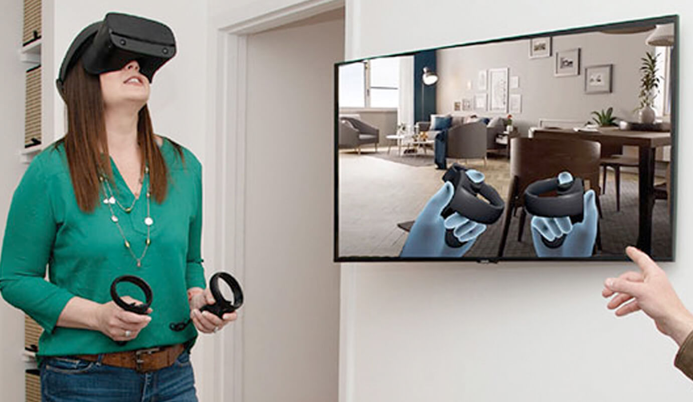 A woman wearing a virtual reality headset and holding controllers stands in a living room. A TV on the wall shows her VR view with animated hands holding controllers in a virtual room.