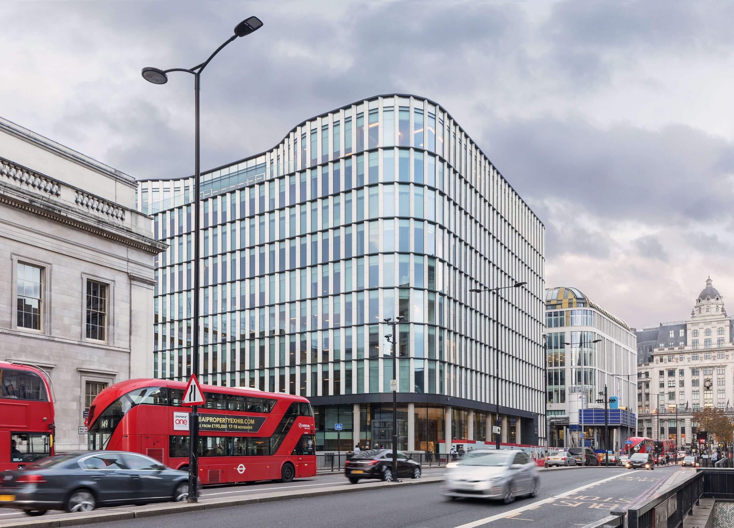 Street view of a modern glass office building with distinctive curved edges, surrounded by traffic including a red double-decker bus. The overcast sky looms over this architects marvel, where BIM principles blend classic and contemporary architectural elements seamlessly.