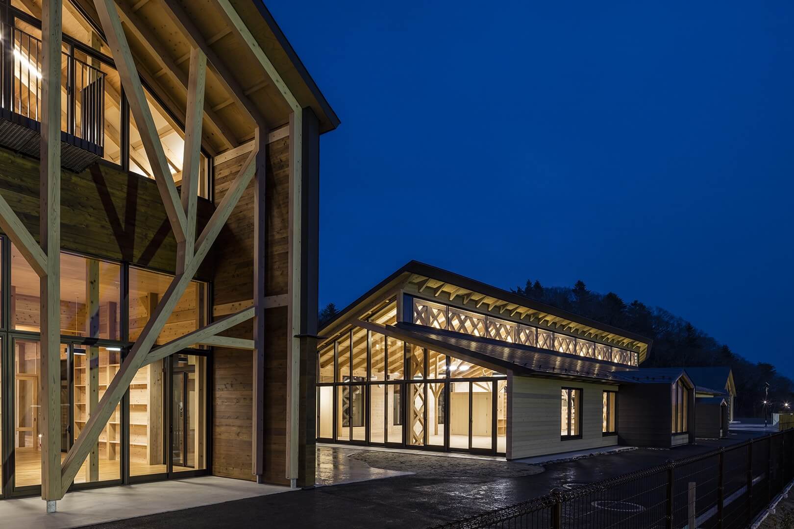 A modern wooden building complex lit from within at night showcases its large windows and exposed beams. Designed using advanced CAD tools, the architecture features angular roofs and a visible interior, all set against a dark blue evening sky.