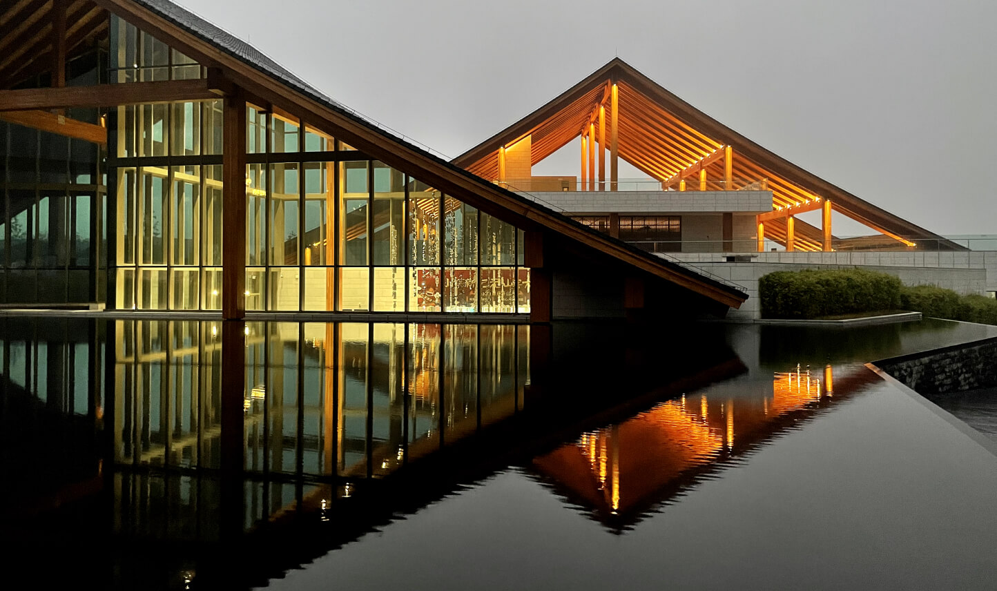 A modern building with sharp, angular roofs is reflected in a still body of water. Created by a visionary architect using BIM technology, the structure is illuminated with warm, glowing lights that contrast against the overcast sky, crafting a serene and harmonious scene.