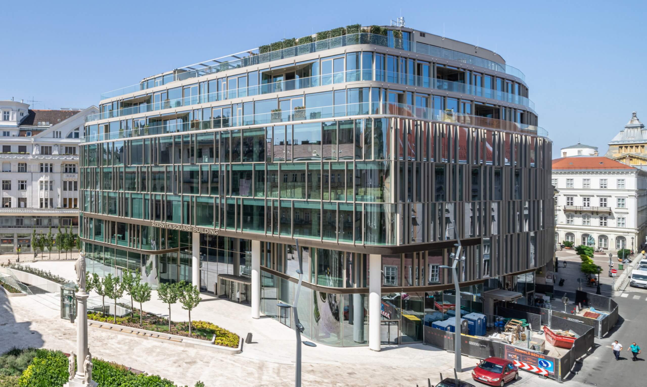 A modern glass-walled building, designed by a renowned architect, boasts multiple stories and a sleek curved design in an urban setting. Surrounding streets are bustling with pedestrians and cars, while adjacent structures feature classic architectural styles.