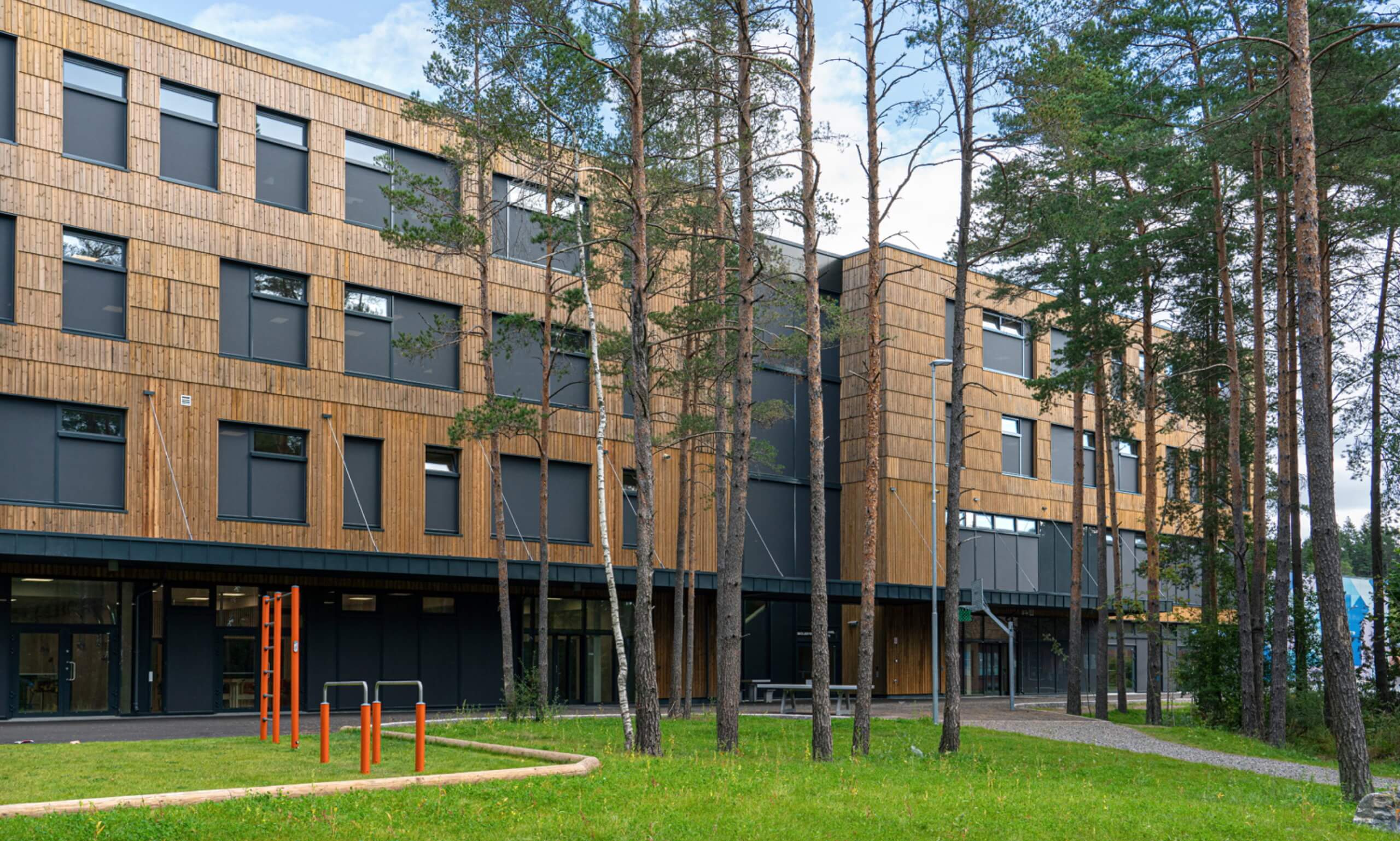 A modern building with a wooden facade, crafted by skilled architects, is surrounded by tall pine trees. In the foreground, theres a grassy area with colorful outdoor exercise equipment. The structure has multiple windows and appears to have three stories.
