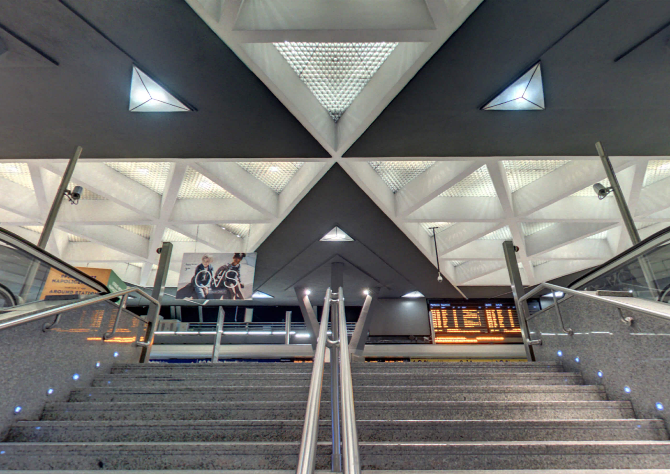View from the bottom of a modern staircase in a train station. The ceiling, a CAD masterpiece, boasts geometric patterns and triangular lights. Below, a digital signboard and a poster with cyclists are visible. The scene showcases Archicads sleek, architectural design.
