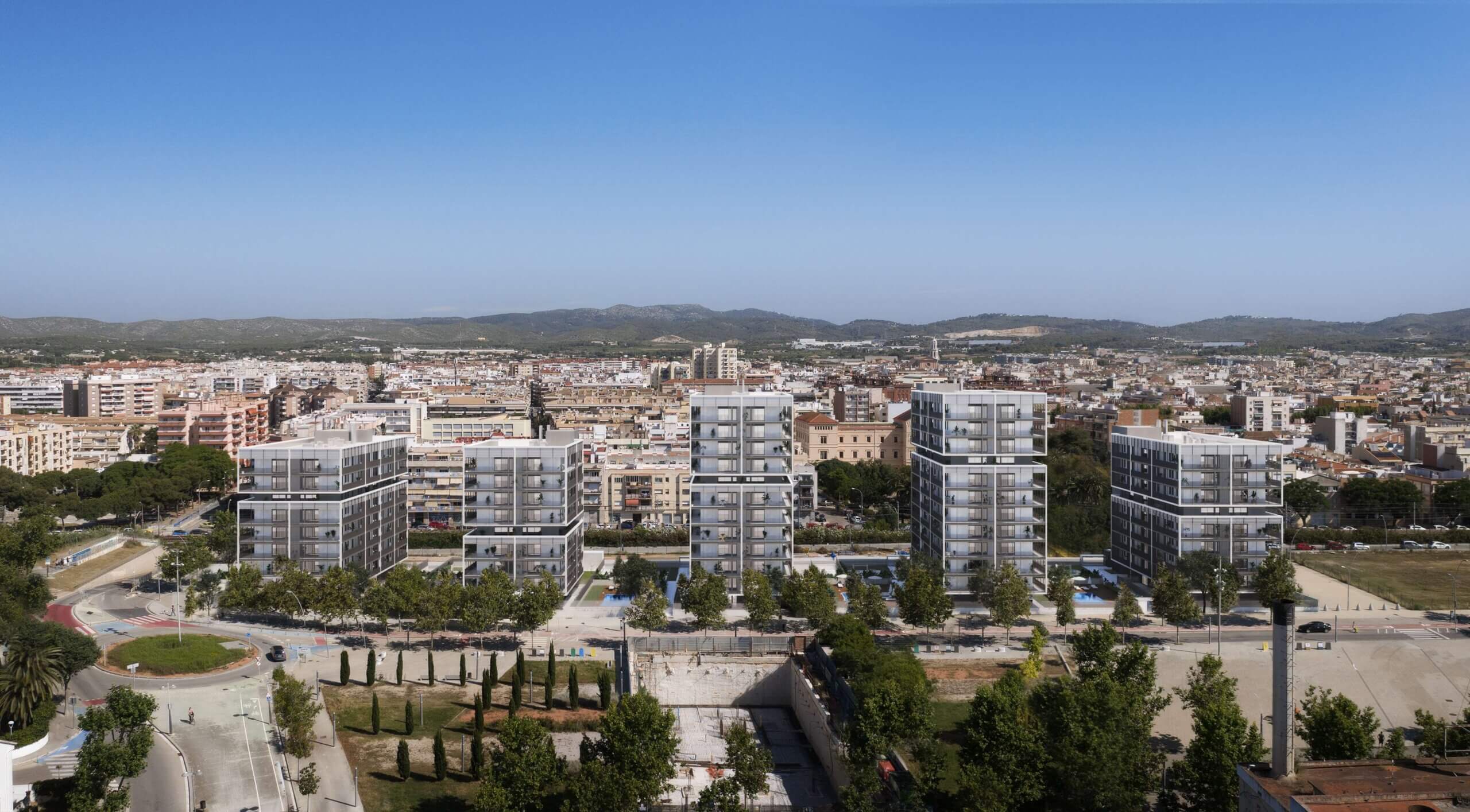 Aerial view of a cityscape with modern mid-rise apartment buildings in the foreground, tree-lined streets, and a sprawling urban area with mountains in the background under a clear blue sky.