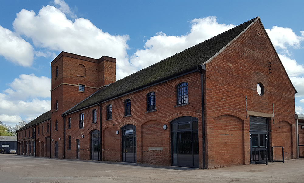 A historic red-brick industrial building with tall arched windows and a gabled roof stands under a partly cloudy blue sky, showcasing the architectural brilliance of its era. The prominent brick tower boasts small square openings, while the foreground features a paved area.