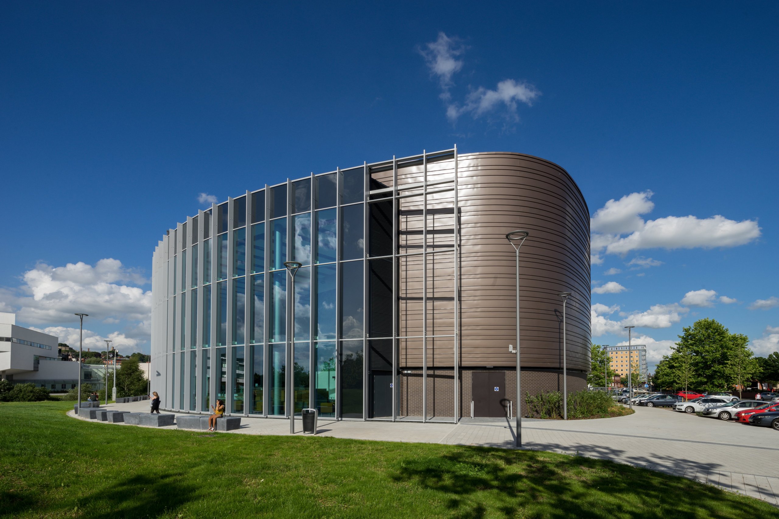This modern building, envisioned by a skilled architect, boasts a curved facade with large glass windows and brown metal paneling against a backdrop of scattered clouds in the blue sky. It is gracefully situated amidst a lawn, pathways, and a few scattered trees.