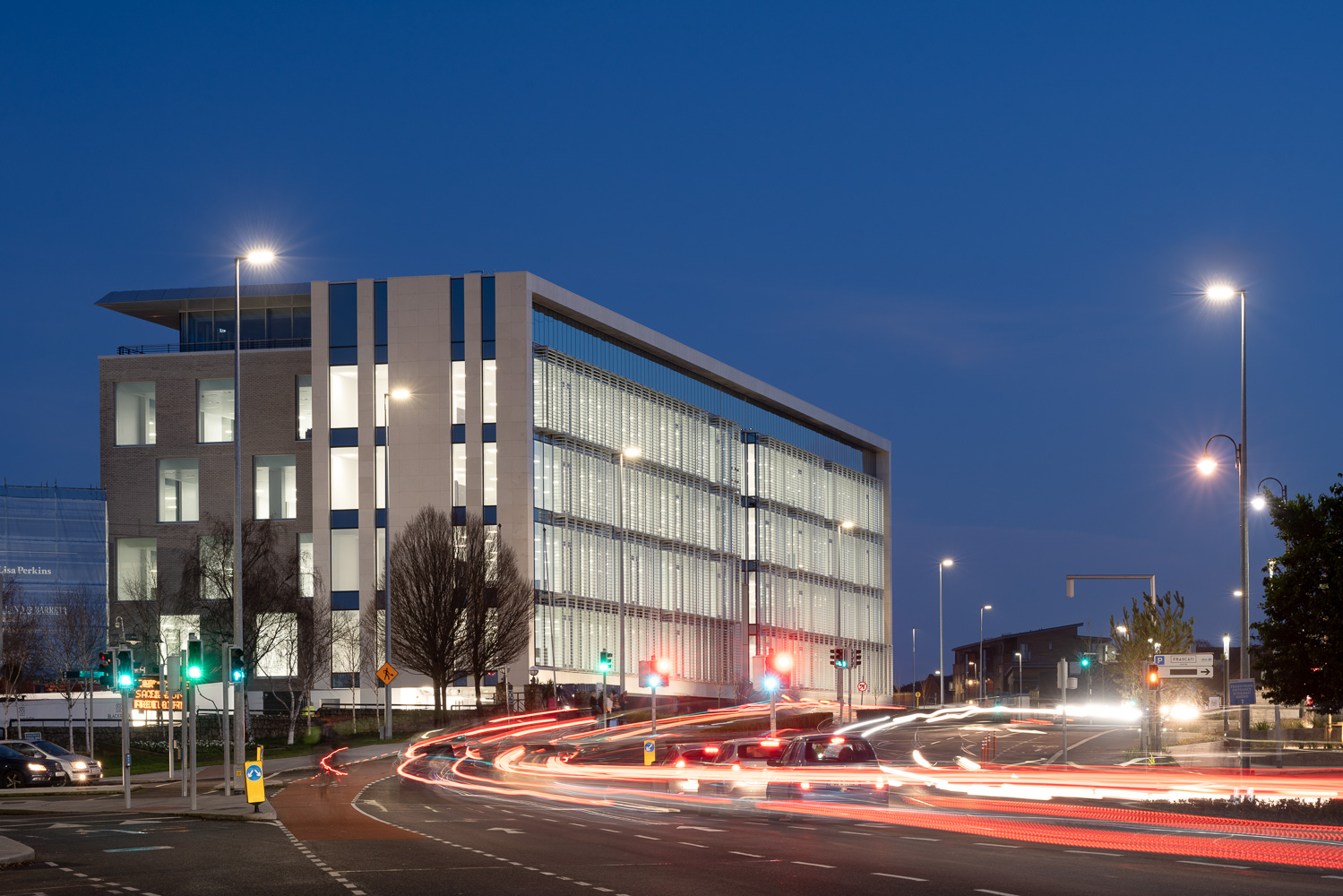 A modern, illuminated office building with large glass windows stands prominently at twilight, embodying the innovation of Graphisoft. The sky is a deep blue, and streaks of car lights create dynamic trails on the road in front, highlighting a busy intersection.