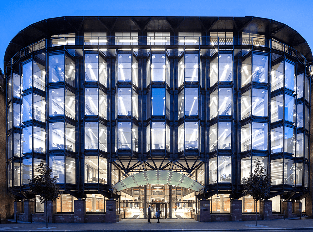 A modern glass building with a grid-like facade glows from within, showcasing its symmetrical, BIM-inspired design. The entrance stands out, framed by two small trees. The sky is a clear blue, indicating early evening.