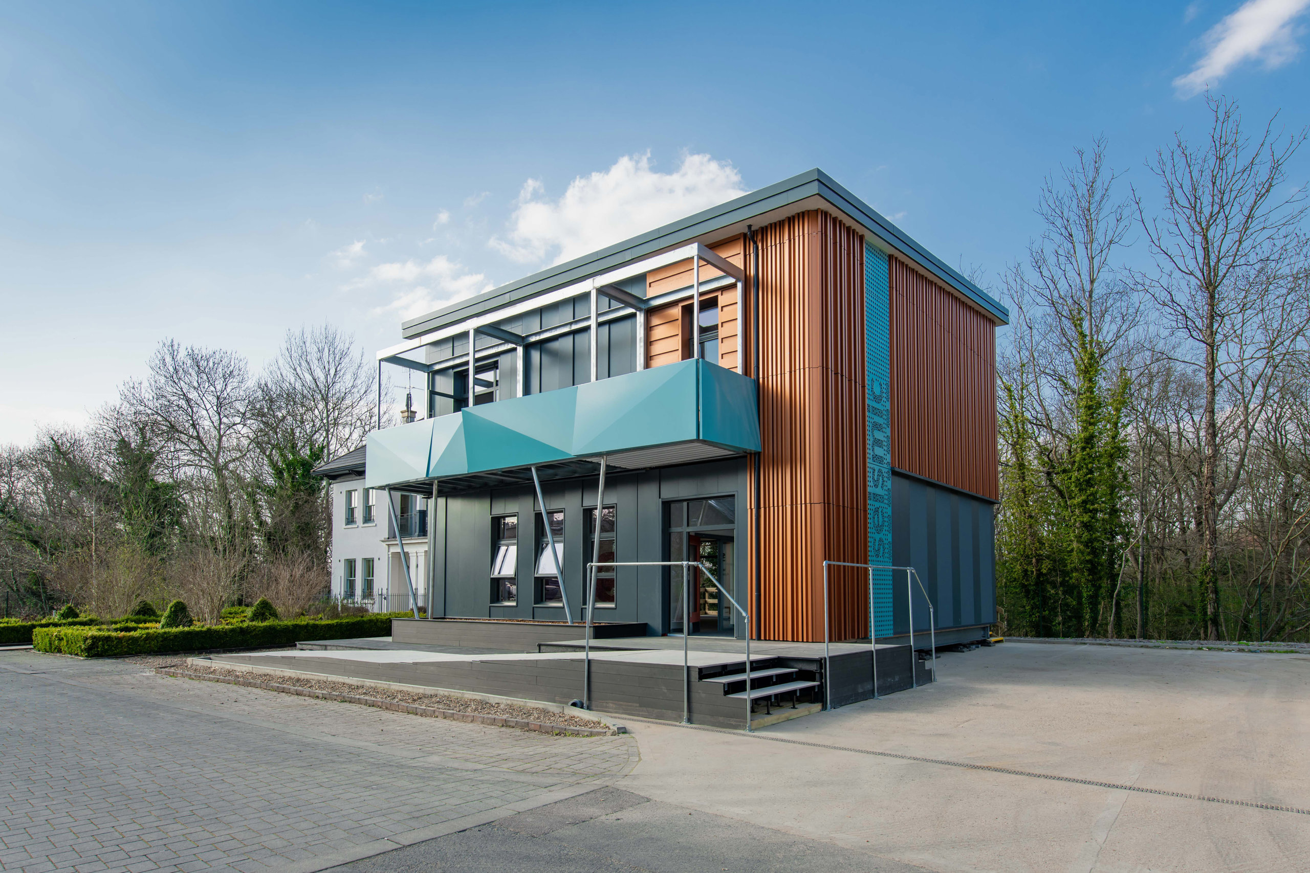 Modern two-story building with orange and blue accents, featuring large windows and geometric design. Surrounded by trees and shrubs, the structure stands on a paved area under a clear blue sky, its design seamlessly brought to life through Archicads cutting-edge capabilities.