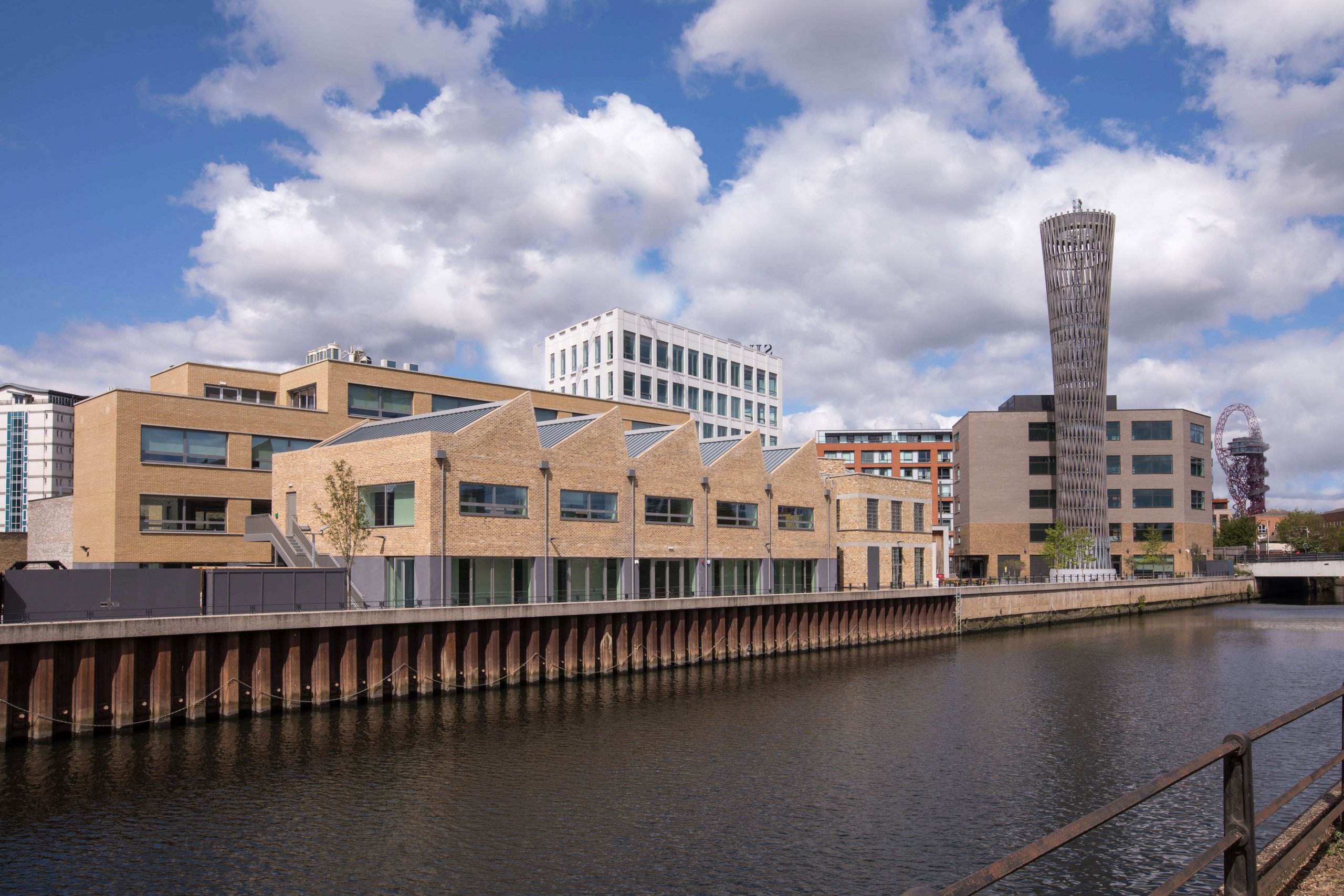 View of modern architecture along a canal, showcasing light brown brick buildings with angular roofs and a tall cylindrical tower. The scene is reminiscent of Graphisofts BIMx models, with reflections glistening in the water under a cloudy blue sky.