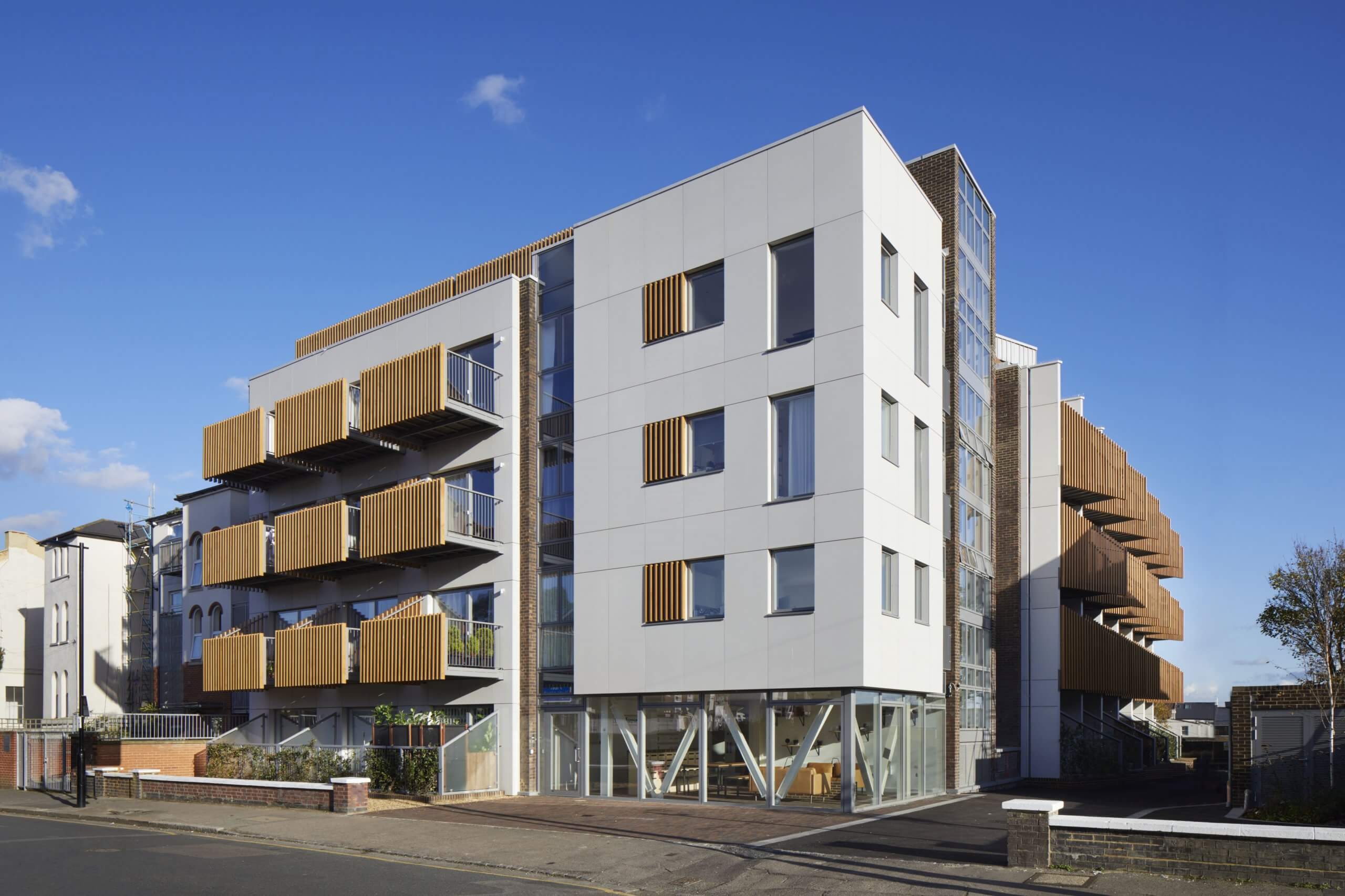 A modern, multi-story apartment building with a sleek design crafted by a visionary architect. It features white and wooden-paneled exteriors, with balconies on each level. The structure has large windows and stands against a clear blue sky, meticulously planned using Archicad software.