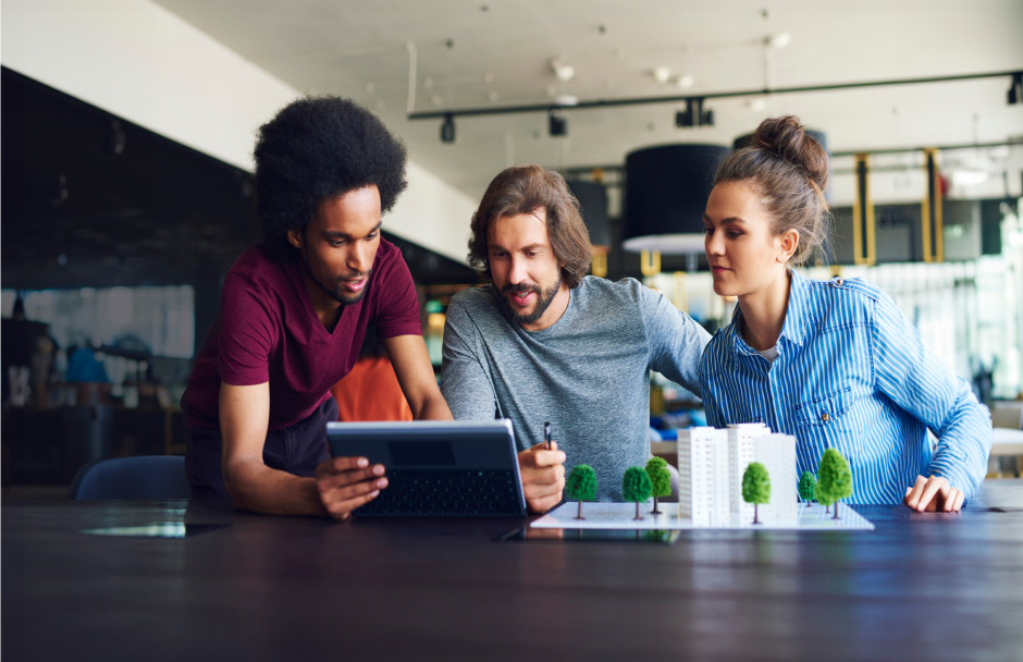 Three architects gather around a table in a modern, well-lit office, discussing and reviewing a tablet. They examine small architectural models of buildings, likely utilizing Archicad or BIM software to enhance their collaborative design process.
