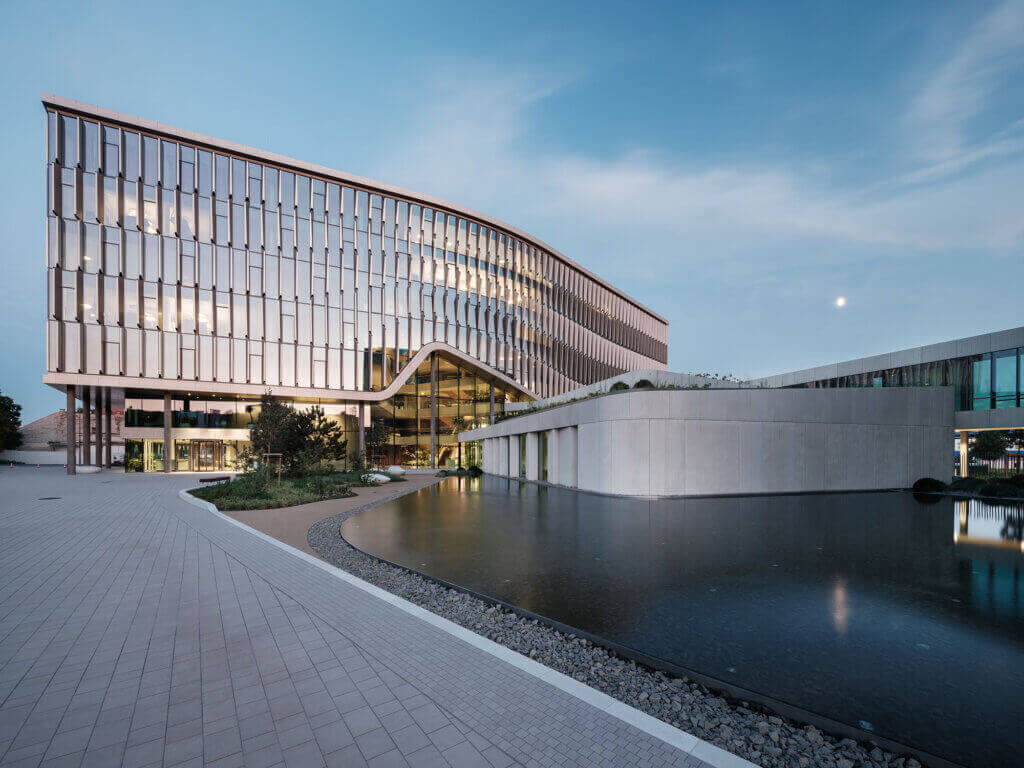Modern glass-fronted building with curved lines, large windows reflecting the sky, and a landscaped area featuring a smooth pond beside a white, angular structure at sunset.