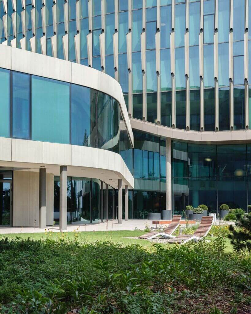 Modern glass building with curved architecture and large windows, surrounded by green lawn and garden. Several lounge chairs are arranged on the grass, creating a relaxing outdoor space in front of the building.