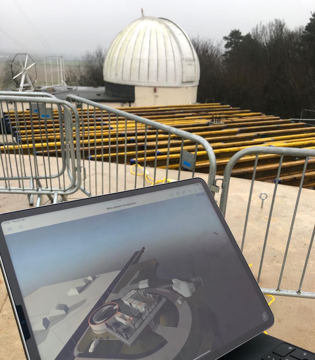Anotherkind Architecture Graphisoft BIMx – Ein Tablet mit einem 3D-Modell wird im Aussenbereich in der Naehe von Metallabsperrungen gehalten, im nebligen Hintergrund sind eine grosse weisse Observatoriumskuppel und gelbe Tragbalken zu sehen.