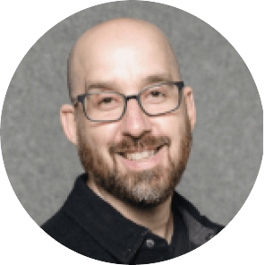 A smiling, bald man with glasses, a trimmed beard, and mustache, wearing a dark collared shirt, posed in front of a plain gray background.