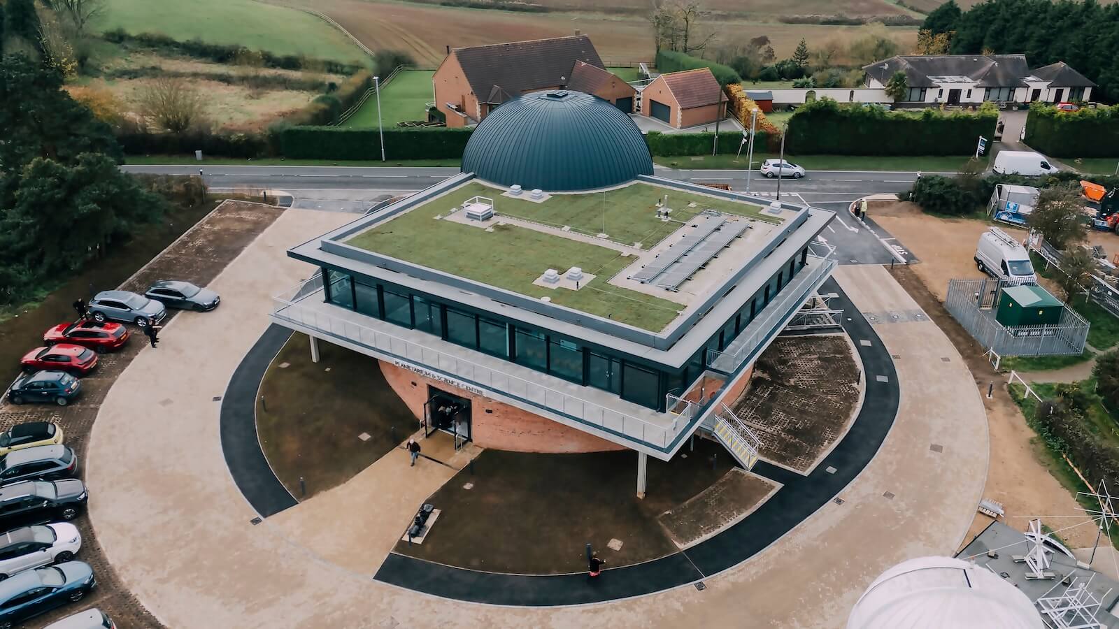Aerial view of Anotherkind Architects' Sherwood Observatory, an adaptive reuse project designed with Graphisoft Archicad. The building has a grass-covered roof and a large black dome, surrounded by pathways, parked cars, and a few houses in the background. Photo credit: G F Tomlinson