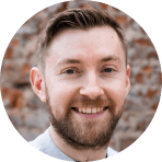 A smiling man with short brown hair and a beard, wearing a light-colored shirt, stands in front of a blurred brick wall background, embodying the spirit of collaborative BIM in his professional demeanor.