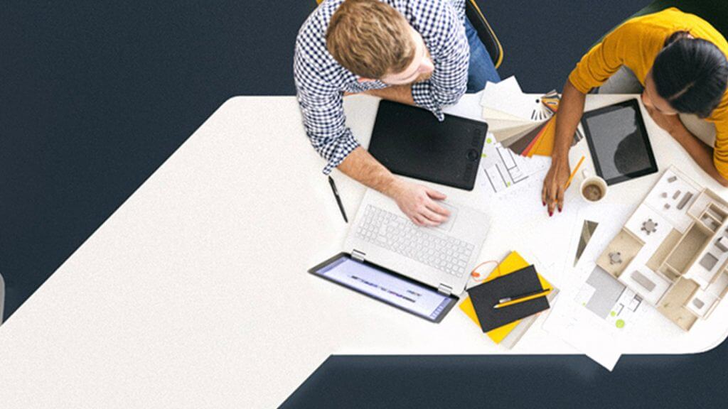 Dos personas sentadas frente a frente en un escritorio blanco, trabajando con un ordenador portátil, una tableta, papeles, un cuaderno, lápices y un modelo arquitectónico, visto desde arriba.