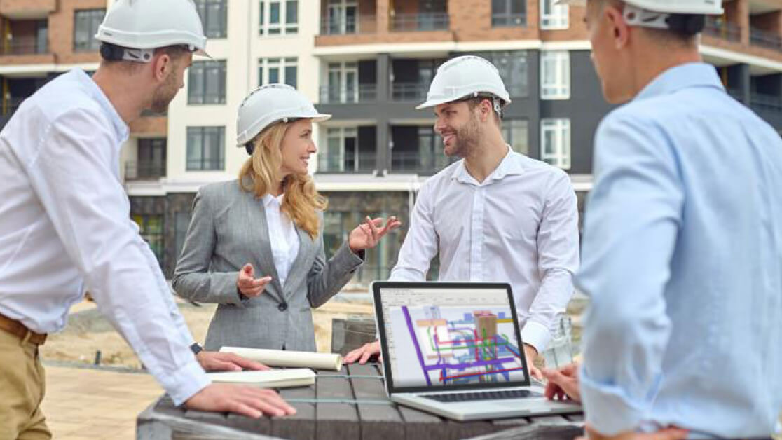 Vier Personen in Schutzhelmen und Geschäftskleidung stehen auf einer Baustelle und besprechen Pläne. Auf dem Tisch vor ihnen steht ein Laptop, auf dem ein Gebäudeentwurf zu sehen ist. Im Hintergrund sind Wohnhäuser zu sehen.
