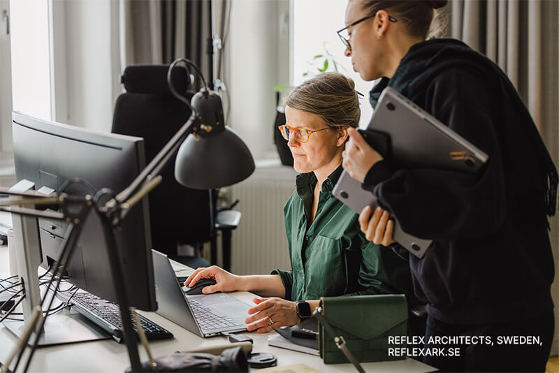 In einem hellen Büro arbeiten zwei Personen an einem Schreibtisch mit Computern. Die eine sitzt, tippt und schaut auf einen Monitor, während die andere daneben steht und einen Laptop hält. Auf dem Schreibtisch liegen Büromaterialien.