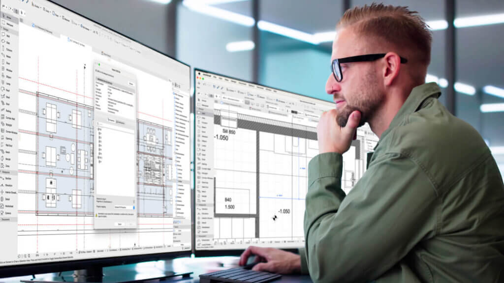 Ein Mann mit Brille und grüner Jacke sitzt an einem Schreibtisch und blickt konzentriert auf zwei große Computermonitore, auf denen architektonische oder technische Planungssoftware mit Gebäudegrundrissen angezeigt wird.