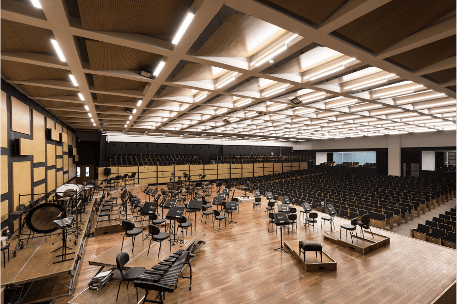 A spacious, modern concert hall with an empty wooden stage set with music stands and chairs, percussion instruments to the left, and rows of empty black seats facing the stage under a grid-patterned ceiling.