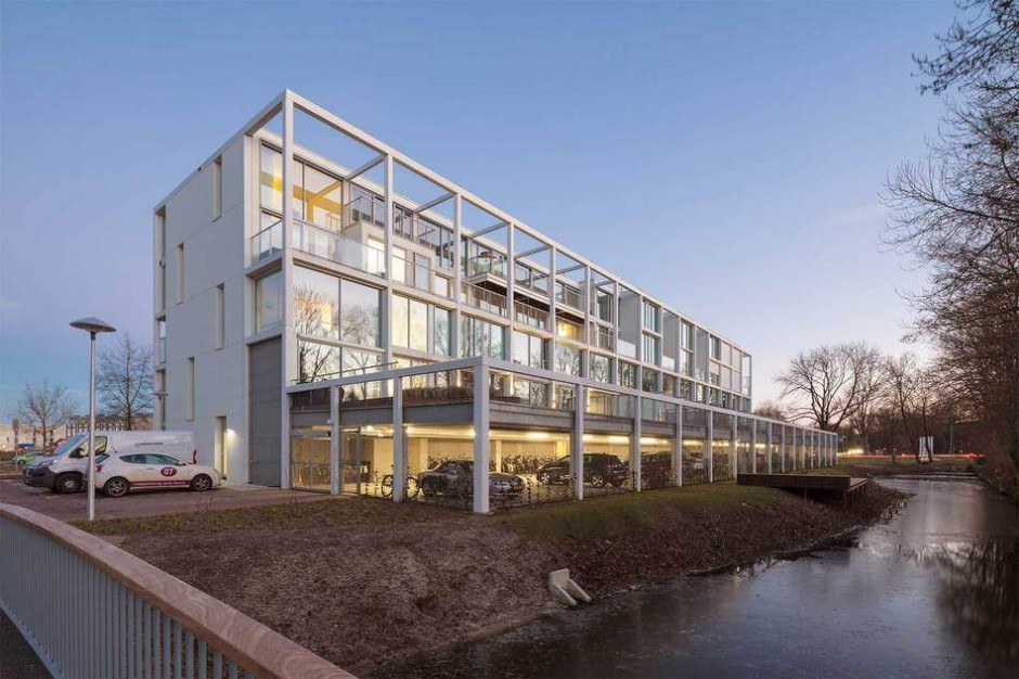 A modern four-story building with large glass windows, visible bicycles parked underneath, and a few cars nearby, situated next to a narrow canal and surrounded by bare trees at dusk.