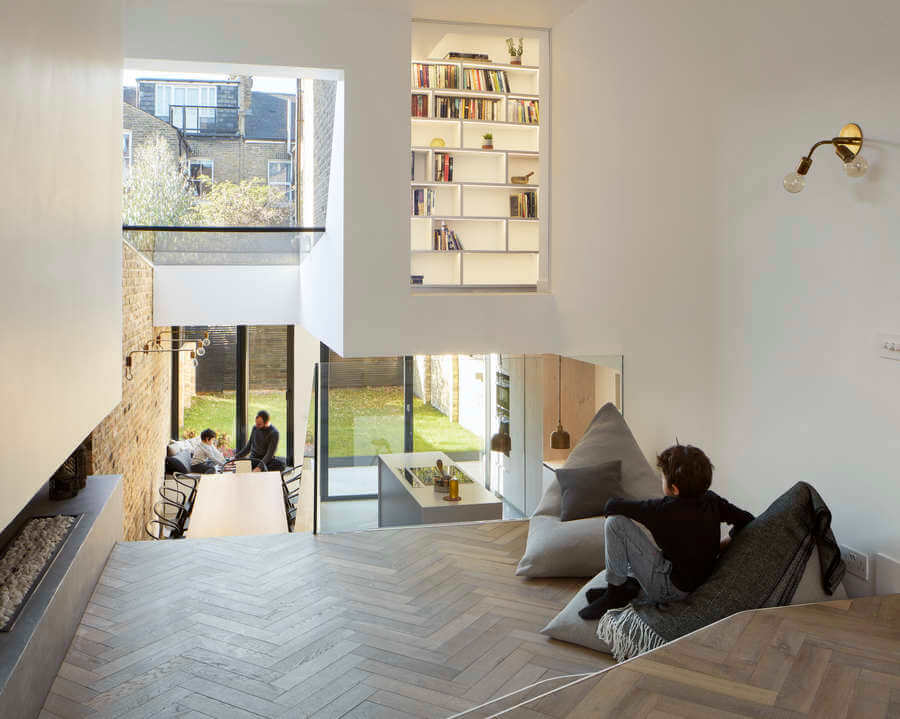 A modern split-level home interior with wooden floors, a boy sitting on a bean bag, built-in bookshelves, large windows, and people sitting at a dining table in the garden outside.