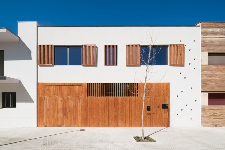 A modern building with a white facade and wooden accents, including window shutters, a large garage door, and mailbox covers. The upper windows have wooden slats, and a small leafless tree stands in front on the sidewalk.