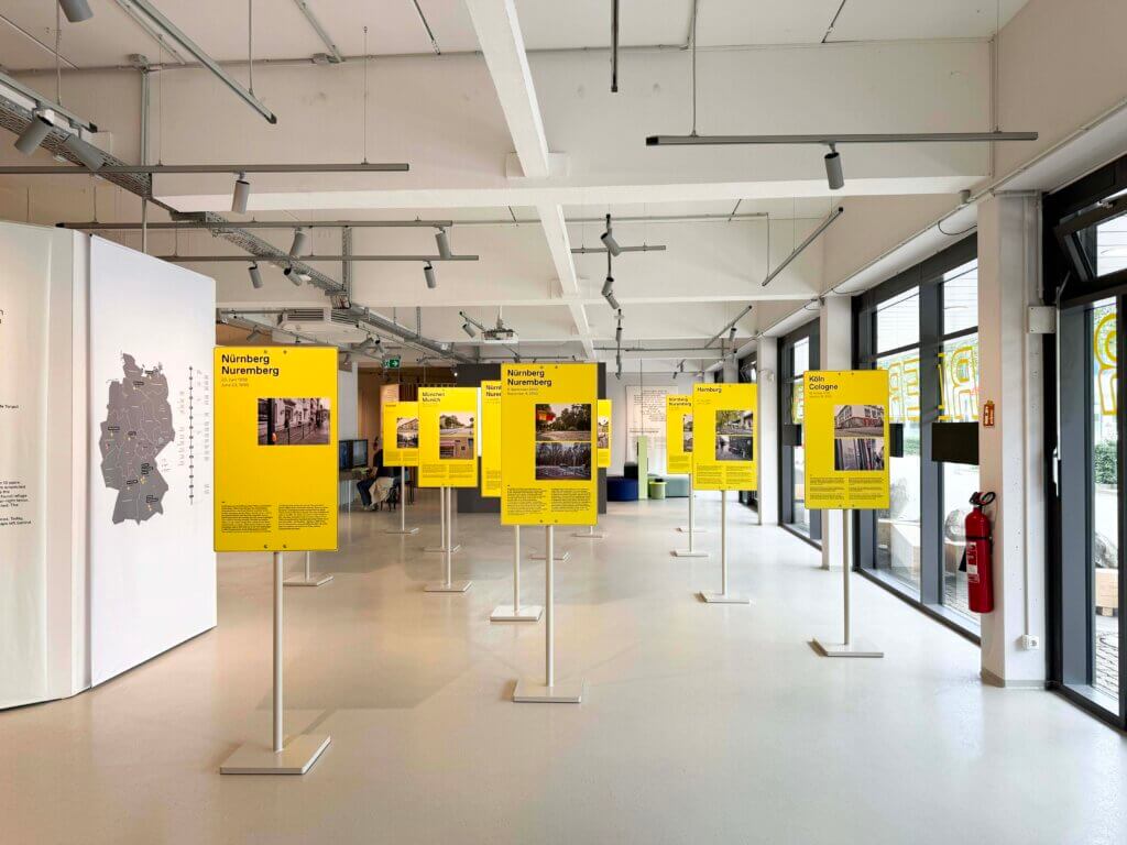 Ein moderner Ausstellungsraum mit großen Fenstern zeigt mehrere gelbe Informationstafeln auf Ständern. Der Raum ist hell, mit einer Landkarte an der linken Wand und einem Feuerlöscher neben dem Eingang.