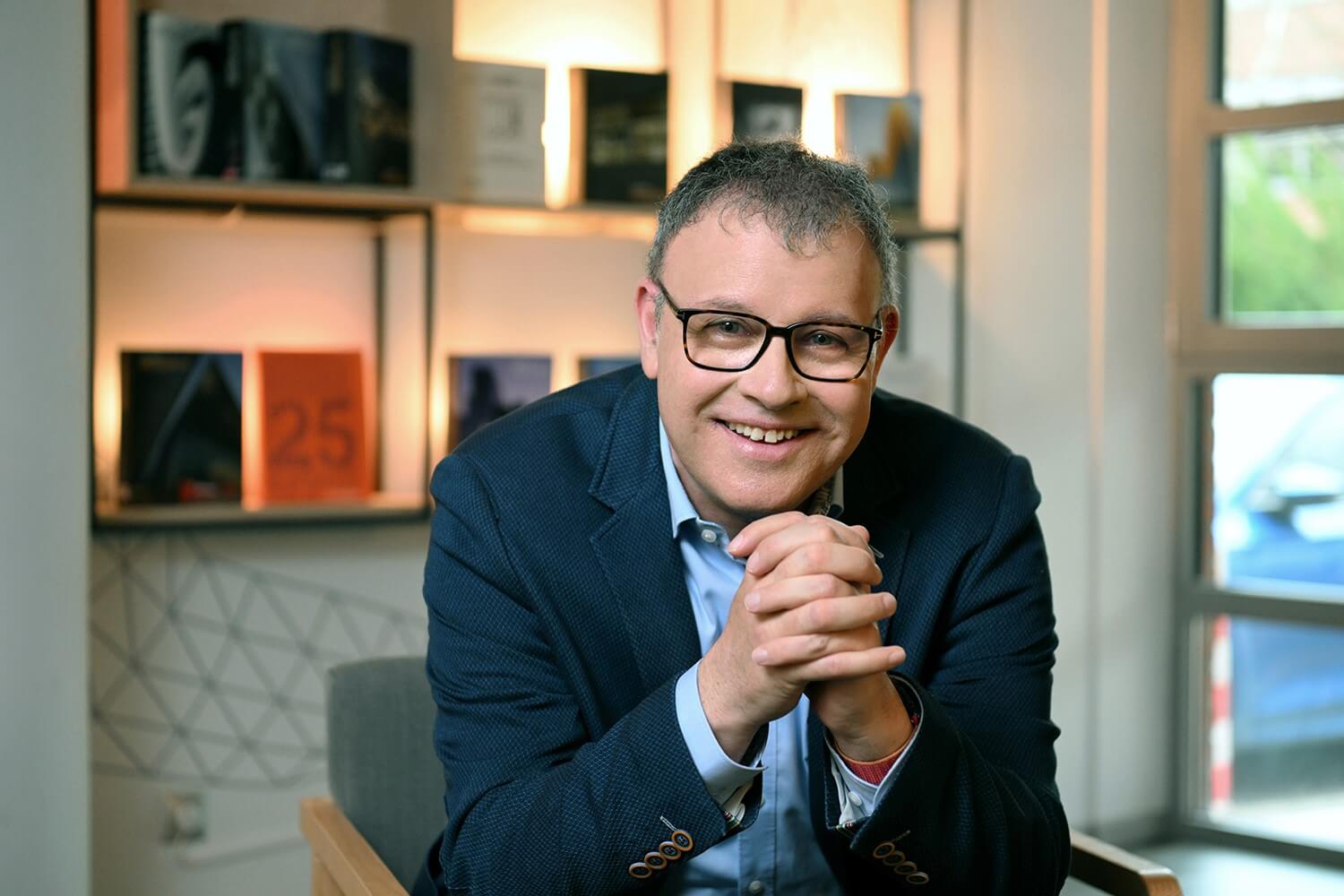Ein lächelnder Mann mit Brille, der einen blauen Blazer und ein helles Hemd trägt, sitzt im Haus und hat die Hände gefaltet. Im Hintergrund sind Bücher und gedämpftes Licht in Regalen zu sehen.